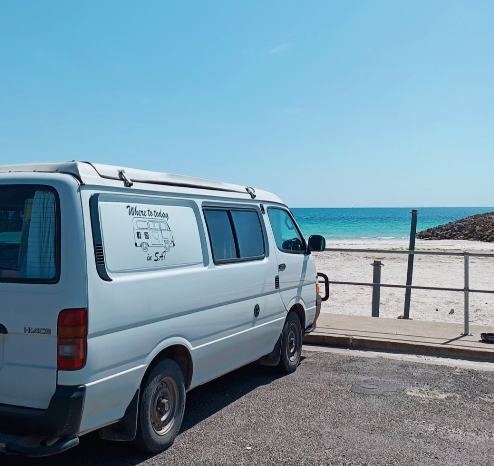 Trevor, Colleen's white hi-top campervan, parked beside a white-sand beach with clear turquoise water beyond.