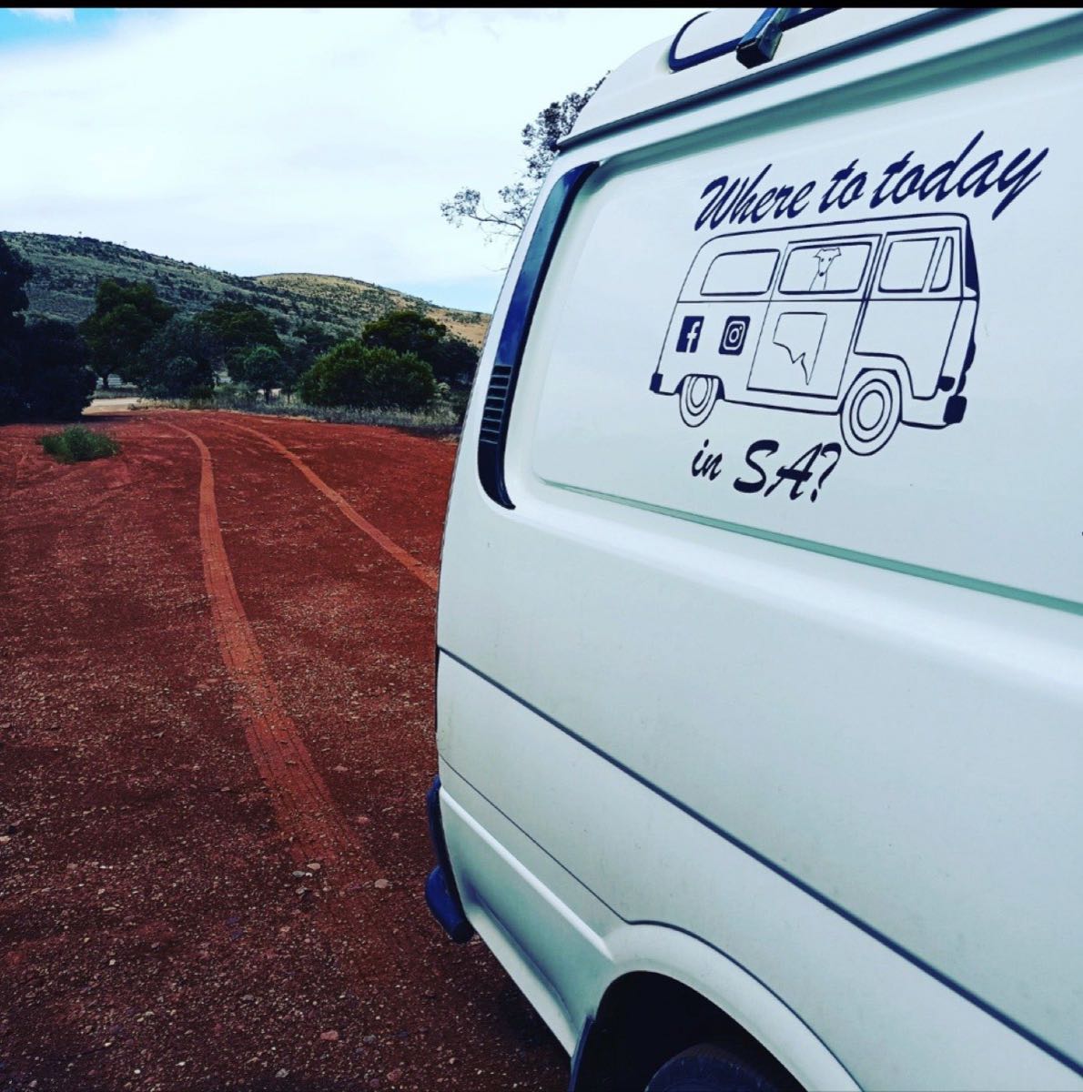 Colleen's white campervan Trevor on a red dirt track, a 'Where to today in SA?' decal on the rear panel and low hills beyond.